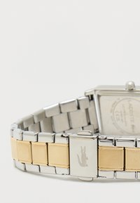 Hõbe- ja kuldtooni metallist rihmaga kell ruudukujulise näo ning nähtava logoga lukul, mis on kombineeritud läikivate ja mati viimistlusega.