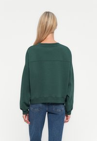 Woman with long blonde hair wearing a dark green sweatshirt and blue jeans, facing away against a plain white background.