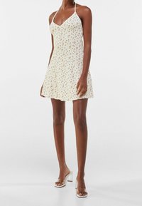 Kvinna iförd en vit halterneck-miniklänning med små blommönster, tillsammans med genomskinliga högklackade sandaler.