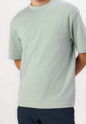 Mand iført en lysegrøn kortærmet t-shirt og mørke bukser med hænderne bag ryggen.
