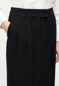 Μαύρο παντελόνι ψηλής μέσης με ραφές και πλαϊνές τσέπες, συνδυασμένο με λευκό πουκάμισο μακρυμάνικο, με το ένα χέρι να ξεκουράζεται στην τσέπη.