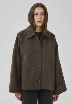 Ženska s ravnimi rjavimi lasmi, oblečena v ohlapno temno rjavo jakno z velikim ovratnikom in sprednjimi žepi ter črne kavbojke.