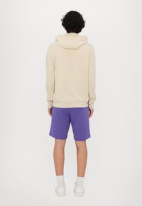 Felpa beige con un cappuccio ampio, abbinata a pantaloni corti viola. L'outfit è realizzato in un tessuto morbido, progettato per un comfort informale.