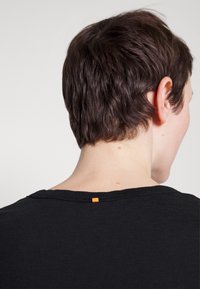 Černé tričko s kulatým výstřihem, na zadní straně s oranžovou nášivkou. Tkanina vypadá měkce a má mírnou strukturu.