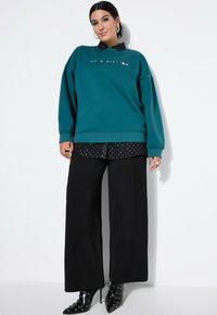Felpa teal con decorazioni colorate, indossata sopra una camicia nera a pois, abbinata a pantaloni neri a gamba larga e stivaletti con borchie.