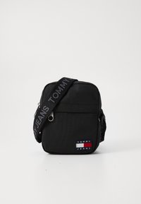 Svart rektangulær crossbody-veske laget av nylon. Har en stoffrem med "TOMMY JEANS" tekst og en liten logopatch med blå, rød og hvite detaljer.