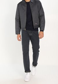 Grå bomberjacka med dragkedja, bärs över en marinblå skjorta. Mörka jeans och vita sneakers fulländar outfiten och betonar en avslappnad stil.