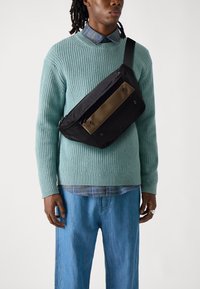 Black and brown crossbody bag made of nylon, featuring a rectangular shape, snap buttons, and a shoulder strap, worn over a green sweater.
