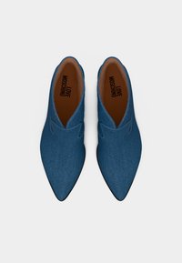 Ζευγάρι μπλε τζιν παπούτσια τύπου slip-on με μυτερή μύτη, με την επιγραφή «LOVE MOSCHINO» στις καφέ εσωτερικές σόλες, εμφανιζόμενα από πάνω σε λευκό φόντο.