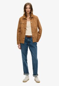 Brun bomuldsjakke med knaplukning, to frontlommer og krave, båret over en beige top og blå jeans, suppleret med hvide sneakers.