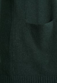 Mörkgrön stickad tröja med en mjuk textur, ribbad nederkant och detalj med ficka fram. Materialet verkar lätt och varmt.