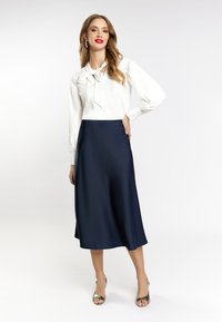 Hvid bluse med sløjfedetalje, ballonsleeves og en marineblå satin-nederdel, elegant silhuet, suppleret af snørebåndssandaler.
