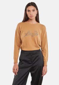 Maglione color cammello con vestibilità rilassata, maniche lunghe e dettaglio di testo argentato luccicante sul davanti. Abbinato a pantaloni neri.