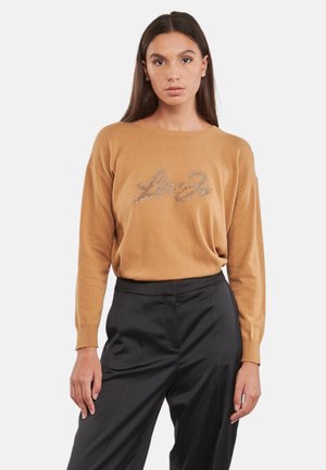 Maglione color cammello con vestibilità rilassata, maniche lunghe e dettaglio di testo argentato luccicante sul davanti. Abbinato a pantaloni neri.