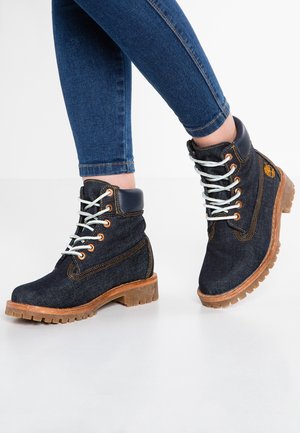 Persona che indossa stivali stringati in denim blu scuro con cuciture color cuoio e suole robuste marroni, abbinati a jeans aderenti blu.