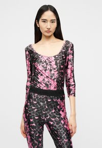 Νεαρή γυναίκα που φοράει μπλούζα με μακριά μανίκια και παντελόνι με ροζ και μαύρο σχέδιο σε σχήμα λέπια, στέκεται μπροστά σε απλό φωτεινό φόντο.