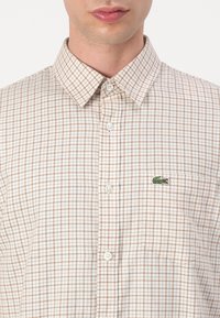 Primer plano de un hombre que lleva una camisa de cuadros beige y marrón con botones y un logo de cocodrilo verde en el bolsillo del pecho.