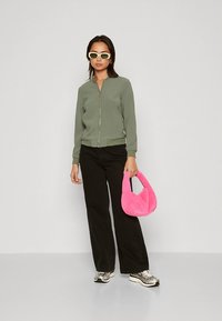 Olive green bomber jacket with a zipper, paired with black wide-leg pants. Accessorized with a fuzzy pink handbag and silver sneakers.
