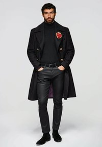 Schwarzer Wollmantel mit goldenen Knöpfen, schwarzer Rollkragenpullover, Lederhose und schwarze Stiefel. Auffällige rote Herz-Anstecknadel am Mantel. Lila Futter sichtbar.