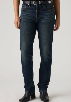 Mørkeblå jeans med lige ben, båret med et sort bælte og sorte ankelstøvler med hæl, vist fra taljen til fødderne mod en ensfarvet baggrund.