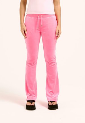 Femme portant un pantalon en velours rose vif et des talons compensés ouverts noirs, debout, les mains détendues le long du corps.