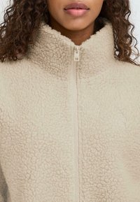 Beež fleece'ist lukuga jope kõrge krae, tekstuurse kangaga ja peene logodetailiga. Omab tavapärast lukustust.