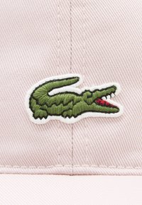 Rožinė beisbolo kepurė su žaliu siuvinėtu krokodilo logotipu ir raudonu akcentu ant struktūrizuotos audinio fono.