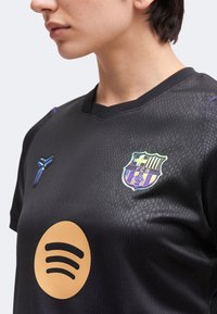 Άτομο με μαύρη ποδοσφαιρική φανέλα της FC Barcelona με το έμβλημα της ομάδας, το λογότυπο του Spotify και ένα γαλαζοπράσινο έμβλημα στο στήθος.