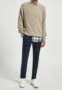 Beżowy sweter w prążki nałożony na granatową koszulę w kratę na guziki, zestawiony z granatowymi spodniami i białymi tenisówkami. Wygląda na swobodny i warstwowy.