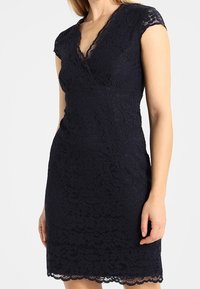 Marinblå spetklänning med korta kapparmarar, V-ringning, figursydd silhuett, scalloped spetskant, detaljerat blommönster och texturerad yta.