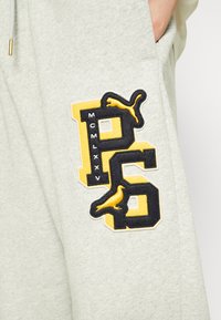 Lys grå sweathose med et stort sort og gult broderet logo med "P" og "6," samt teksten "MCMLXXV" sammen med et fugledesign.