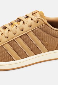 Brun Adidas Superstar sneaker med tre läderränder, textilmaterial, snörning och avvit sula på en vit bakgrund.