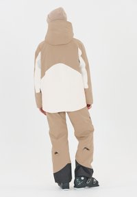 Giacca da sci e pantaloni con blocchi di colore beige e bianco, materiale impermeabile, design con cappuccio, con accenti neri sui pantaloni e stivali da sci.