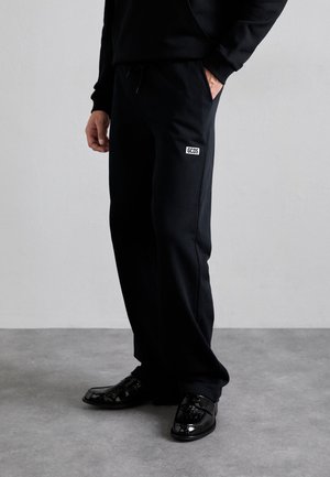 Man draagt zwarte joggingbroek met een GCDS-logo op de dij, gecombineerd met glimmende zwarte nette schoenen, staand op een grijze vloer.