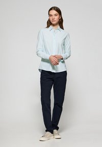 Camicia button-up blu chiaro in un tessuto morbido, abbinata a pantaloni blu navy scuri e sneakers beige. La camicia presenta un colletto classico e una vestibilità slim.