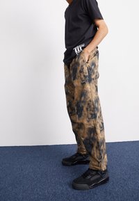 Lösa, batikmönstrade byxor i nyanser av brunt och svart, med sidofickor, kombinerade med svarta sneakers och en svart skjorta.