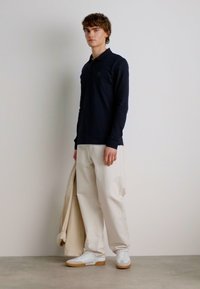 Marineblauwe polo met lange mouwen, romige wijde broek, witte sneakers en een lichte jas, tegen een neutrale achtergrond. Eenvoudig ontwerp en pasvorm.