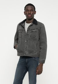 Jachetă din denim gri cu un guler din blană întunecată, cu închidere prin nasturi în față, două buzunare pe piept și croială clasică, asortată cu blugi bleumarin închiși.