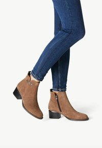Botines de ante marrón con un bajo tacón cuadrado y cremallera lateral, combinados con unos jeans ajustados de un azul oscuro, mostrando una silueta elegante.