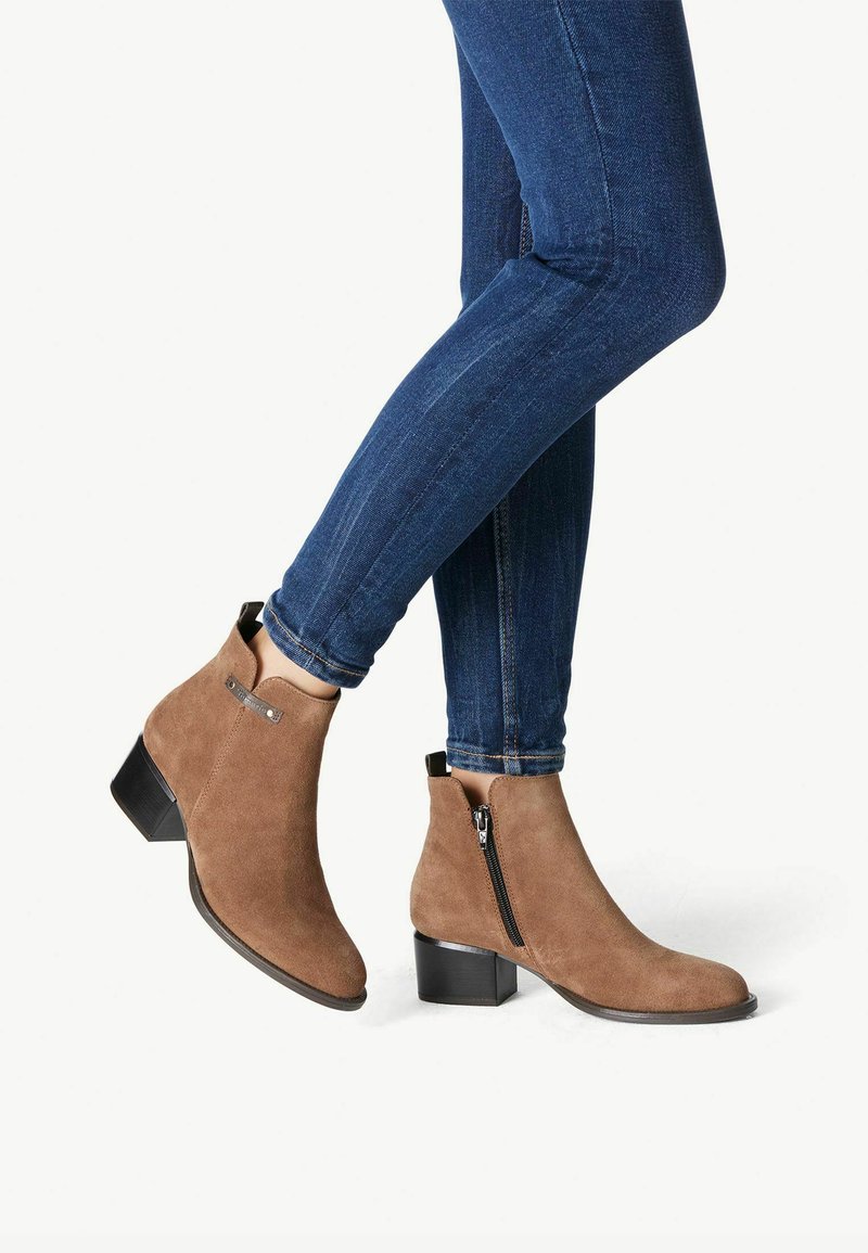 Botines de ante marrón con un bajo tacón cuadrado y cremallera lateral, combinados con unos jeans ajustados de un azul oscuro, mostrando una silueta elegante.