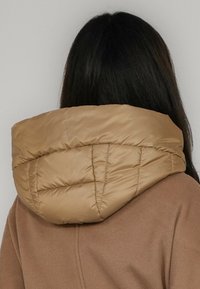 Brązowa puchowa kurtka z dużym pikowanym kapturem. Materiał wydaje się gładki i błyszczący, a przeszycia podkreślają teksturę.