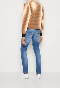 Person wearing blue slim-fit jeans, white sneakers, and an orange and white striped long-sleeve shirt, standing against a plain white background.