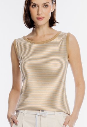 Kobieta w bezrękawniku w beżowo-białe paski z beżowym wykończeniem, w połączeniu z jasnymi spodniami z białym paskiem sznurowanym.