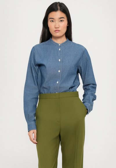 Giovane donna con lunghi capelli scuri che indossa una camicia di denim blu con bottoni, infilata in pantaloni a vita alta di colore verde oliva, con le mani nelle tasche.