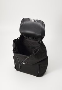Bolsa negra de nailon con una solapa de cuero texturizado, cierre de cuerda y acentos de herrajes plateados. El interior está forrado y es espacioso.