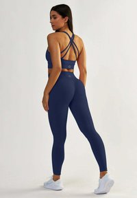 Granatowy stanik sportowy z krzyżującymi się ramiączkami na plecach, w połączeniu z granatowymi legginsami o wysokim stanie. Materiał wydaje się gładki i elastyczny. Białe tenisówki dopełniają wygląd.