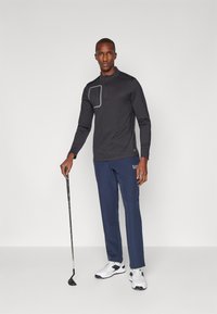Schwarzes Langarmshirt mit halbem Reißverschluss und geometrischem grauem Akzent, kombiniert mit dunkelblauen Hosen und weißen Sportschuhen. Golfschläger in der Hand.