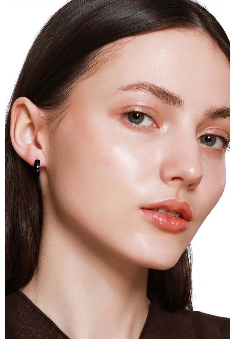 Schwarzes Hohrring, der am linken Ohr getragen wird, mit glatter Textur und minimalistischer Gestaltung. Das Modell zeigt klare Haut und dezentes Makeup.