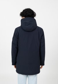 Marineblaue Parka mit Kapuze, mit einer glatten Textur und einem längeren hinteren Saum. Verfügt über Seitentaschen und minimale Nähte für einen schlanken Look.
