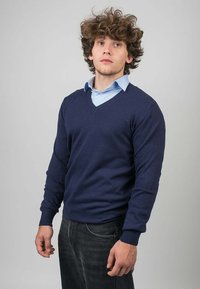 Námořnický svetr s výstřihem do V vyrobený z pleteného materiálu, přiléhavý design s dlouhými rukávy, kombinovaný s světle modrou košilí s límečkem pod ním. Černé džíny.